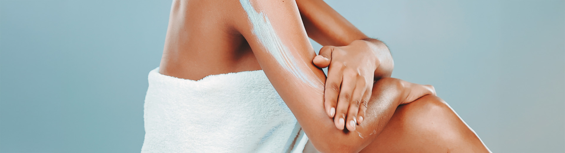A person with a white towel wrapped around them rubbing a skin cream containing Urea on to the dry skin on their arm.