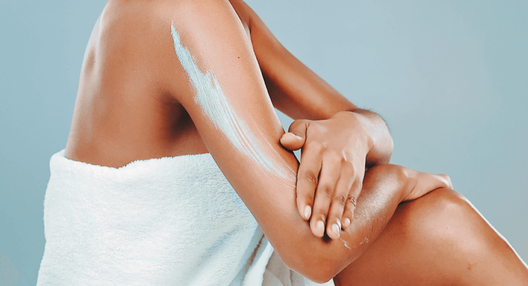 A person with a white towel wrapped around them rubbing a skin cream containing Urea on to the dry skin on their arm.
